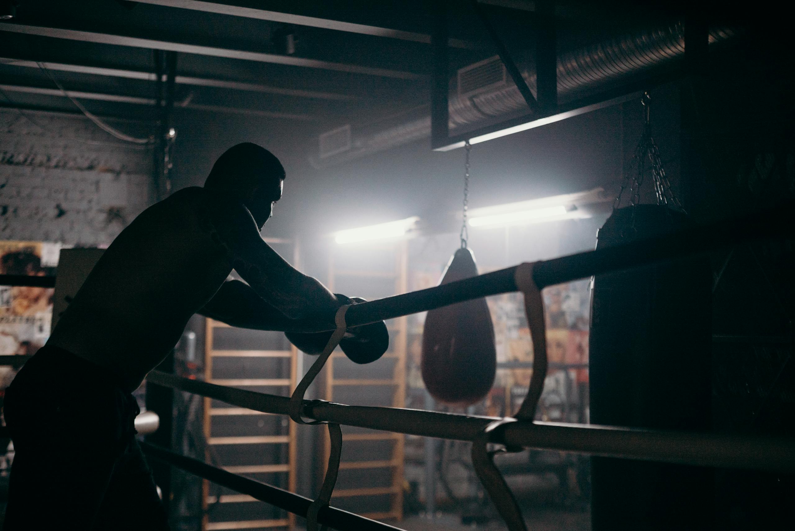 Moody silhouette of a boxer leaning in a dimly lit gym with punching bag and boxing ring.
